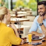 A man and woman sitting across from each other in a coffee shop. They're smiling naturally while making eye contact. Neither is touching the other yet, but both are clearly engaged in the conversation, displaying signs of mutual attraction.